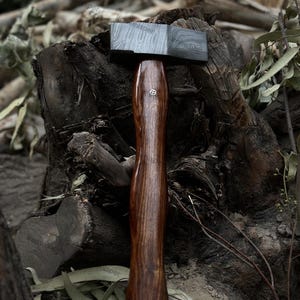 May include: A handcrafted hammer with a dark, patterned steel head and a polished, dark wood handle. The handle features a decorative metal inlay. The hammer is designed for woodworking and metalworking.