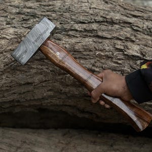May include: A handcrafted hammer with a patterned steel head and a dark wooden handle. The handle has a smooth, polished finish and a decorative inlay. The hammer is held against a textured, natural wood background.