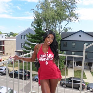 Pode incluir: Um vestido curto vermelho estilo frente única com o texto branco INDIANA HOOSIERS e um logótipo de tridente branco no peito. O vestido tem uma silhueta justa com detalhes de franjas ao longo das costuras laterais e um decote profundo.