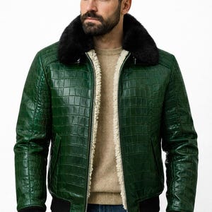 Könnte beinhalten: Grüne Leder-Bomberjacke mit geprägtem Krokodilmuster. Die Jacke hat einen schwarzen Pelzkragen, ein cremefarbenes Innenfutter und schwarze gerippte Bündchen und Saum. Ein silberner Reißverschluss verläuft vorne.