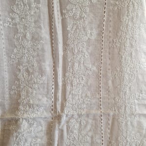 Puede incluir: Tela blanca con intrincado bordado floral. El diseño presenta paneles verticales de flores y hojas bordadas, con delicados detalles calados. La tela parece ligera y adecuada para ropa o decoración del hogar.