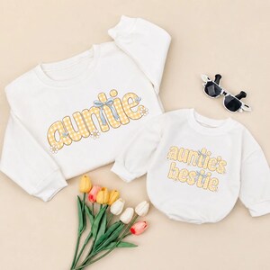 May include: A white long-sleeve sweatshirt with the word auntie in yellow gingham print and a white long-sleeve onesie with the text auntie's bestie in yellow gingham print, both featuring blue bow and white daisy graphics, plus black sunglasses.