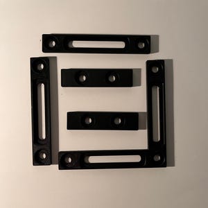 May include: Black metal brackets with slotted holes and circular mounting holes are arranged on a white surface. The brackets are rectangular and L-shaped, designed for structural support and assembly.