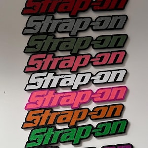 May include: A collection of embroidered patches with the text "Strap-on" in various colors, including blue, red, gray, green, pink, white, orange, and purple. Each patch has a black border and a unique color scheme.