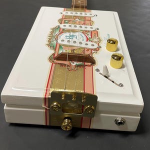 May include: A handmade cigar box guitar with a white body, adorned with gold and red accents. The guitar features a wooden neck, gold tuning knobs, and a decorative label. The design incorporates a bridge, pickups, and a volume control.