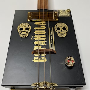 May include: A handmade cigar box guitar with a black body and gold hardware. The guitar features gold skull accents, the text "Hand Made Fine Cigars", and a skull-shaped knob with red eyes. The neck is made of wood.