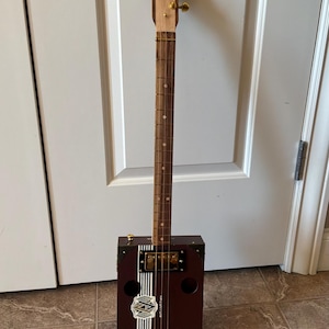 May include: A handmade cigar box guitar with a dark brown rectangular body and a wooden neck. The body features a decorative label and vertical white stripes. The headstock is light-colored wood with gold tuning pegs. The guitar has four strings.