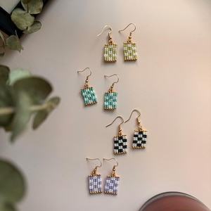 Peut inclure: Plusieurs paires de boucles d'oreilles perlées faites à la main, de couleurs variées : vert, turquoise, noir et blanc, et violet. Chaque boucle d'oreille est de forme rectangulaire avec des crochets dorés. Présentées sur une surface rose clair.