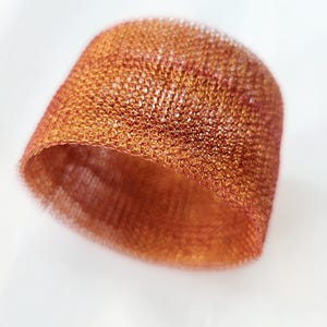 Pode incluir: Um close-up de uma escultura de malha de arame de cor cobre. A malha é tecida em um padrão apertado, criando uma superfície texturizada. A escultura tem a forma de uma tigela ou um chapéu.