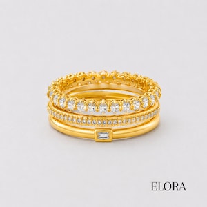 May include: A stack of three gold rings. The top ring features a row of round clear stones. The middle ring is a thin band encrusted with small clear stones. The bottom ring is a smooth gold band with a single rectangular baguette stone.