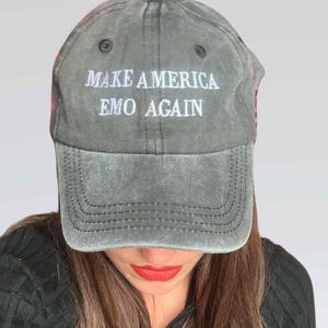 May include: A gray baseball cap with the text "MAKE AMERICA EMO AGAIN" in white. The cap has a curved brim and a stitched design. The person wearing the cap has long brown hair and red lipstick.