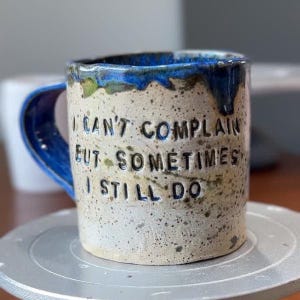 May include: A speckled ceramic mug with a blue glazed rim and handle. The mug features the embossed text I CAN'T COMPLAIN BUT SOMETIMES I STILL DO in dark blue lettering. The body of the mug has a light beige, textured, speckled finish.