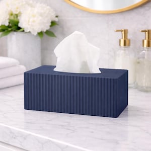 May include: A navy blue rectangular tissue box with a vertical ribbed design. White tissues protrude from the top. The box is on a white marble countertop, with a gold-framed mirror and soap dispensers in the background.