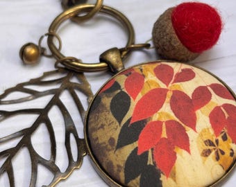 May include: A keychain featuring a round pendant with a fabric insert displaying a red and yellow leaf pattern. The keychain includes a bronze-toned metal leaf charm, a small bell, and a red felted ball set in a brown acorn cap, all attached to a ring.