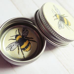 May include: Two small, open, silver-colored tins with lids. Each lid and the inside base of the tins feature a yellow and black bumblebee design. The bumblebee is centered on a light yellow background.