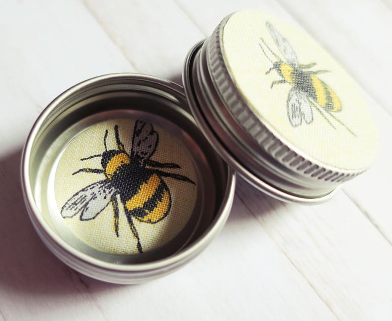 May include: Two small, open, silver-colored tins with lids. Each lid and the inside base of the tins feature a yellow and black bumblebee design. The bumblebee is centered on a light yellow background.
