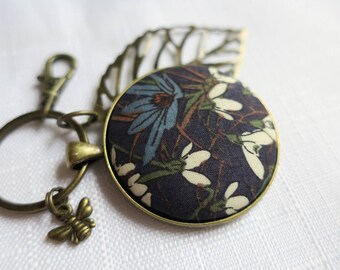 May include: A round keychain featuring a fabric-covered button with a dark blue floral pattern, including white and light blue flowers. The keychain has an antique bronze-colored metal finish, a small bee charm, and a decorative leaf-shaped metal cutout.