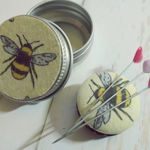 May include: A small, round metal tin with a lid featuring a yellow and black bee design. Beside it, a matching pin cushion with three heart-topped pins. The bee design is repeated on the pin cushion.