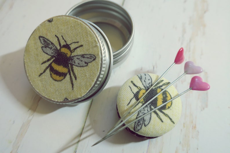May include: A small, round metal tin with a lid featuring a yellow and black bee design. Beside it, a matching pin cushion with three heart-topped pins. The bee design is repeated on the pin cushion.
