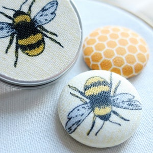 May include: Close-up of three fabric-covered buttons with bee and honeycomb designs. One button is in a silver tin. The bee buttons feature black, yellow, and gray details. The honeycomb button is orange and yellow.
