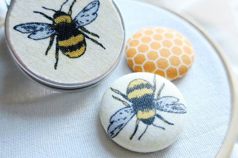 May include: Close-up of three fabric-covered buttons with bee and honeycomb designs. One button is in a silver tin. The bee buttons feature black, yellow, and gray details. The honeycomb button is orange and yellow.