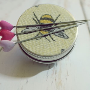 May include: A small, round container with a fabric top featuring a bee illustration. Three heart-shaped pins in shades of pink and purple are placed on top, creating a charming and decorative sewing accessory.