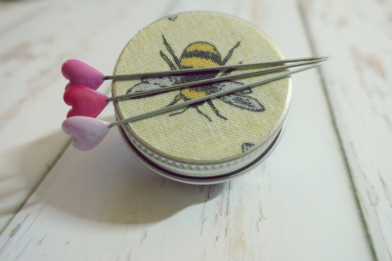 May include: A small, round container with a fabric top featuring a bee illustration. Three heart-shaped pins in shades of pink and purple are placed on top, creating a charming and decorative sewing accessory.
