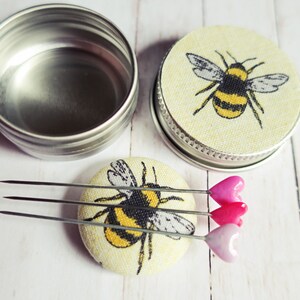May include: A set of sewing pins with heart-shaped heads in shades of pink, resting on a round, fabric-covered pin cushion. The cushion features a yellow and black bumblebee design. A silver tin and a second pin cushion with a bee design are also present.