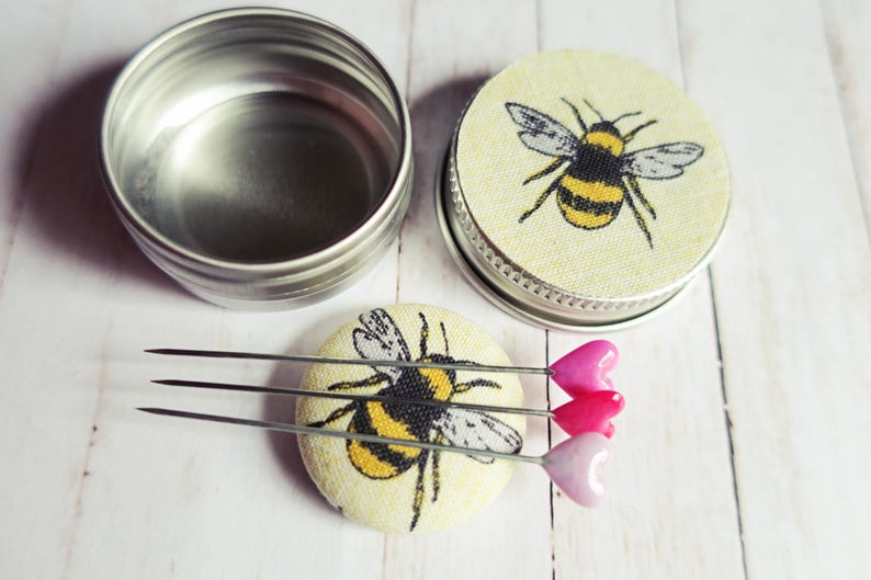 May include: A set of sewing pins with heart-shaped heads in shades of pink, resting on a round, fabric-covered pin cushion. The cushion features a yellow and black bumblebee design. A silver tin and a second pin cushion with a bee design are also present.