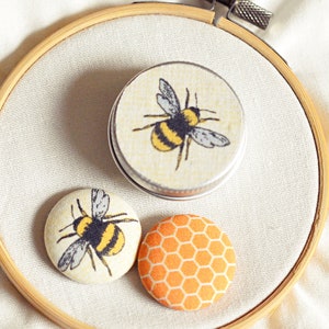 May include: A wooden embroidery hoop holds a small tin and two fabric-covered buttons. The tin and one button feature a yellow and black bee design. The other button has an orange honeycomb pattern.