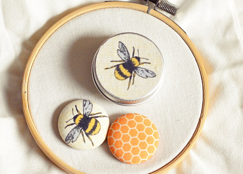 May include: A wooden embroidery hoop holds a small tin and two fabric-covered buttons. The tin and one button feature a yellow and black bee design. The other button has an orange honeycomb pattern.