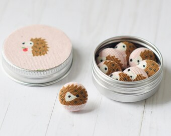 May include: A set of small, round fabric-covered buttons featuring a brown and cream hedgehog design. The buttons are stored in a round silver metal tin, with one additional button placed outside the tin and the lid featuring a hedgehog graphic.