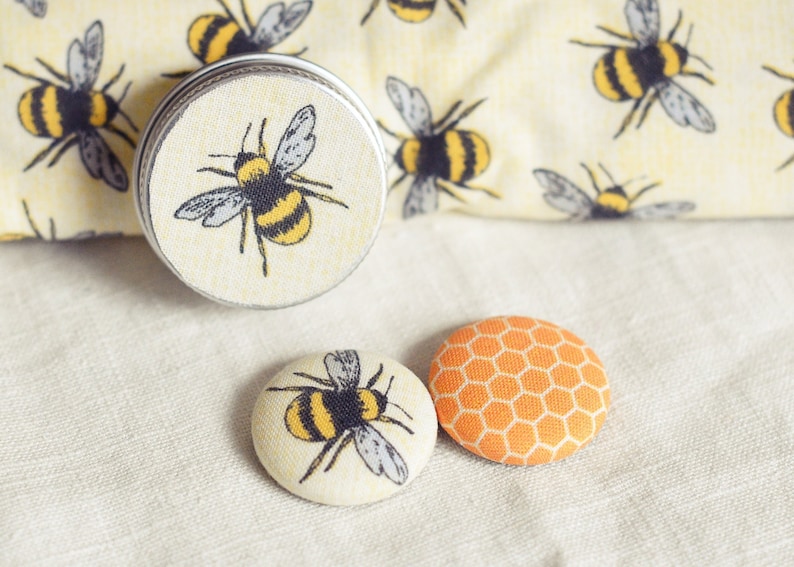 May include: A metal tin and two fabric-covered buttons with bee designs. The tin and one button feature a bee illustration with yellow and black stripes. The other button has an orange honeycomb pattern.