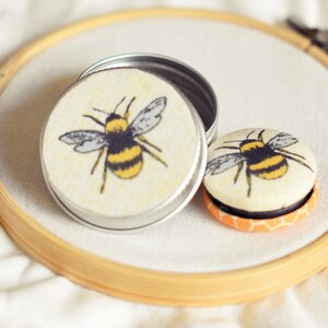 May include: A small, open silver tin and a round button with a bee design. The bee has yellow and black stripes and gray wings. The items are on a white surface, inside a wooden embroidery hoop.