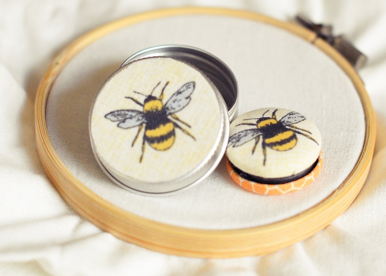 May include: A small, open silver tin and a round button with a bee design. The bee has yellow and black stripes and gray wings. The items are on a white surface, inside a wooden embroidery hoop.