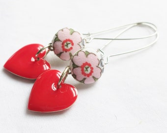 May include: A pair of dangle earrings featuring a shiny red heart-shaped charm suspended from a round fabric-covered bead. The bead displays a light pink floral pattern with green leaves and a red center. The earrings have silver-toned metal hooks.