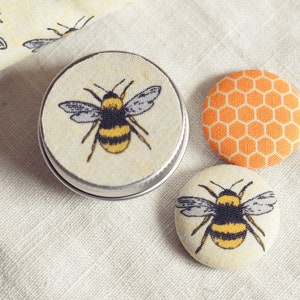 May include: A small, round tin with a bee design, along with two fabric-covered buttons. One button features a honeycomb pattern, and the other has a bee illustration. The items are on a cream-colored fabric.