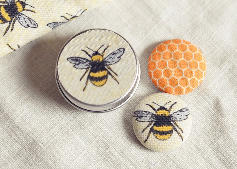 May include: A small, round tin with a bee design, along with two fabric-covered buttons. One button features a honeycomb pattern, and the other has a bee illustration. The items are on a cream-colored fabric.