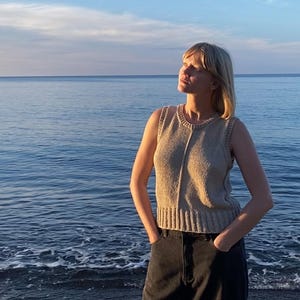Puede incluir: Un top sin mangas de punto beige con dobladillo acanalado y cuello redondo. La persona lleva vaqueros negros y está de pie cerca del mar. El cielo es azul con algunas nubes.