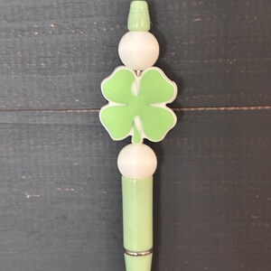 May include: A light green pen with a four-leaf clover design. The pen features a white ball and a light green four-leaf clover charm. The pen is accented with a silver band and a small green top.