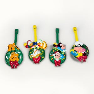 May include: Four plastic luggage tags featuring Disney characters Pluto, Donald Duck, Mickey Mouse, and Goofy. Each tag depicts a character within a green holiday wreath adorned with a red bow and includes a yellow or green plastic attachment strap.