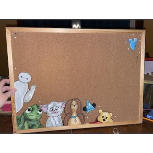 May include: Corkboard with a wooden frame, featuring hand-painted cartoon characters. The characters include Baymax, a green frog, a white creature, a brown-haired dog, a blue wizard hat, and Winnie the Pooh. A blue balloon is painted in the upper right corner.