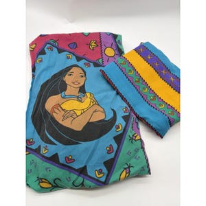 Op de afbeelding: Een gevouwen stoffenset met een Pocahontas-karakterafbeelding op een blauwe achtergrond met een geometrische rand. De set bevat een apart stuk met horizontale banden van gele, paarse en groene patronen, geaccentueerd door kleine bladmotieven.