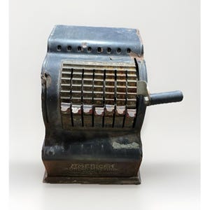 Puede incluir: Una antigua máquina sumadora negra de la marca American, con un asa y varias teclas numéricas. La máquina tiene forma rectangular con una parte superior ligeramente curvada y un asa metálica en el lateral. El texto "AMERICAN" es visible.