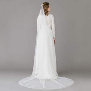 May include: A white wedding dress with long sleeves and a flowing skirt. The dress has a delicate lace bodice and a long, sheer veil. The bride is seen from the back, with her hair styled up. The veil trails on the floor.