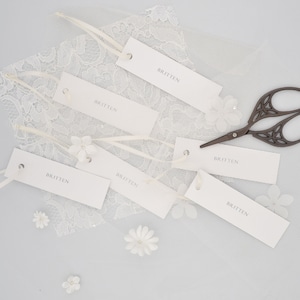 Op de afbeelding: Zes witte rechthoekige labels met het woord "Britten" erop gedrukt. De labels zijn gerangschikt op een wit oppervlak met witte kant en kleine witte bloemen. Een paar zwarte decoratieve scharen bevindt zich ook op het oppervlak.