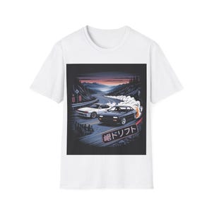 以下が含まれることがあります： 白いTシャツ。山道でドリフトする2台の車のグラフィック。イラストはレトロスタイルで、日本語のテキストが入っています。車は白と黒で、煙のエフェクトがあります。背景には、山の上で夕焼けが見えます。