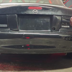 Peut inclure: Vue arrière d'une voiture de sport Mazda MX-5 Miata noire. La voiture a un pare-chocs noir, des feux arrière et le logo Mazda. La zone de la plaque d'immatriculation est noire. Les pneus sont visibles en bas de l'image.