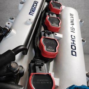 Può includere: Primo piano di un motore di auto con componenti bianchi e bobine di accensione NGK rosse. Il motore presenta la scritta "Mazda" in blu e "DOHC 16-VALVE" in nero. È visibile un tappo dell'olio motore blu.