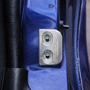 May include: A close-up of a silver metal car door hinge with two bolts, mounted on a blue car door. The hinge has the text "miria solutions" printed on it. The door's black interior is visible.